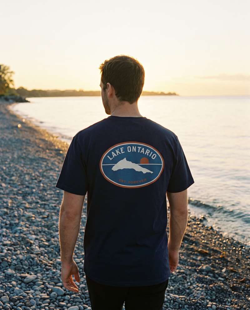 Man walking on a Lake Ontario pebble beach wearing the True Navy tee with the big badge back print