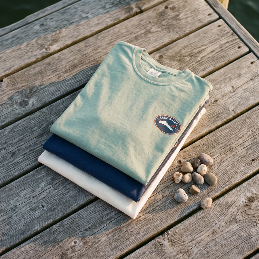 Stack of folded Lake Ontario heavyweight tees in seafoam, true navy and white on dock wood