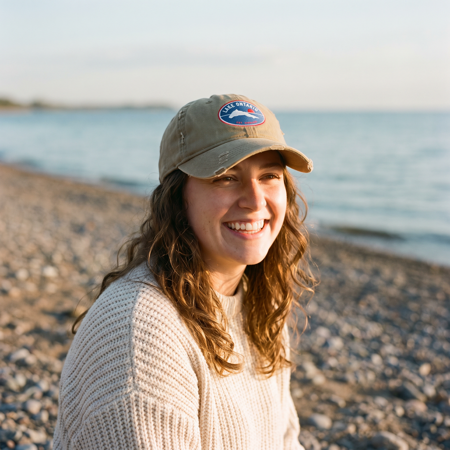 Woman wearing the khaki Lake Ontario badge cap on a pebble beach